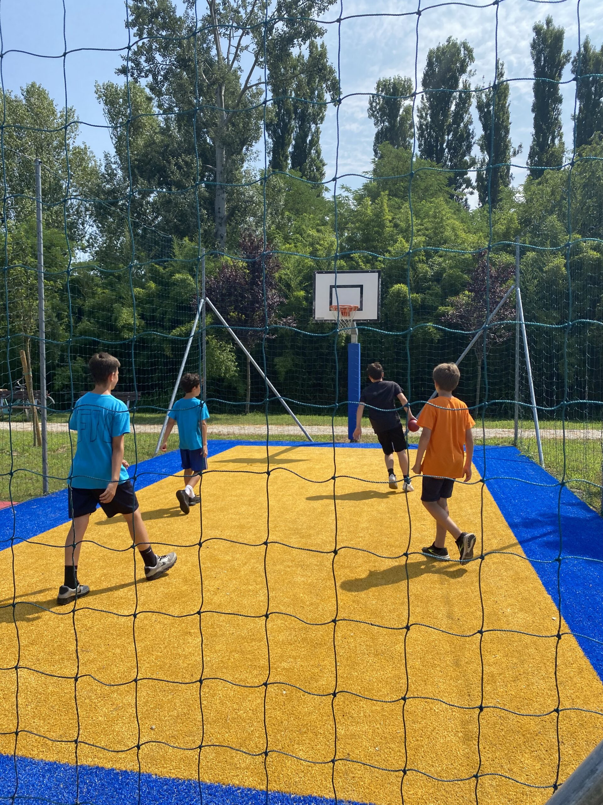 campo sportivo delimitato da una rete lungo tutto il suo perimetro. C'è un canestro e la superficie è un tappeto morbido giallo con una cornice blu a contrasto.
